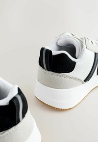 Balti ir balti sportiniai bateliai su zomšiniais akcentais, turintys masyvų baltą pado, tekstūruotą audinį ir dvi juodas juostas ant kulno.