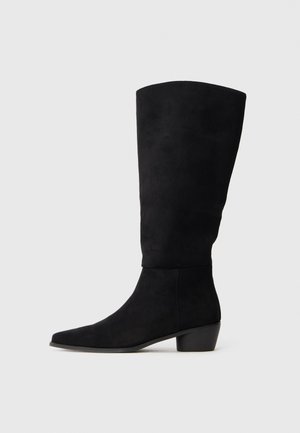 Bota alta de ante negra hasta la rodilla con tacón bajo y punta afilada, mostrada en vista lateral sobre un fondo gris claro liso.