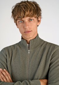 Maglione grigio con zip, caratterizzato da un motivo testurizzato e colletto rialzato, con zip silver e maniche lunghe. Modello con le braccia incrociate.