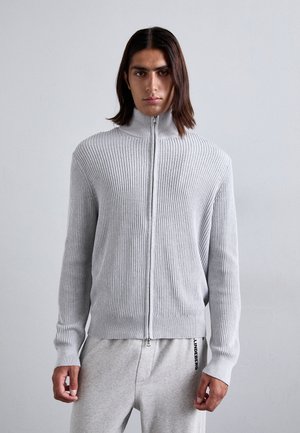 Lys grå ribbestrikket zip-sweater med høy krage og lange ermer, sammen med grå joggebukser med logo på venstre ben.