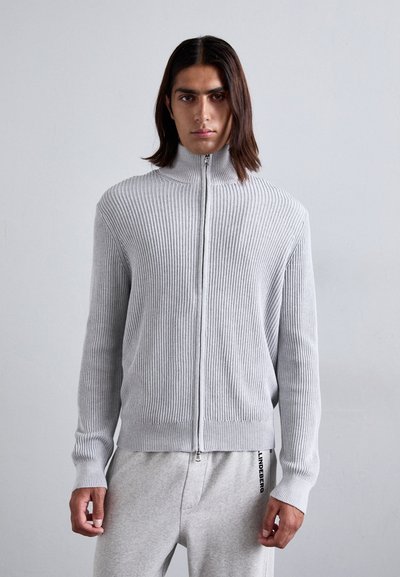 Sweat à zip gris clair avec côtes, col haut et manches longues, associé à un jogging gris avec un logo sur la jambe gauche.