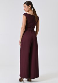 Burgunderfarbener Jumpsuit mit tailliertem Oberteil, weitem Bein, Rundhalsausschnitt und Reißverschluss im Rücken. Besitzt eine taillierte Taille mit goldenen Knopfakzenten.