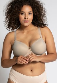 Bra imbottito beige con spalline regolabili, tessuto liscio e un piccolo fiocco decorativo al centro. Il modello indossa slip a vita bassa coordinati.
