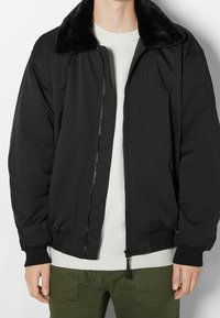 Svart bomberjacka med krage i fuskpäls, dragkedja, ribbade manschetter och avskärning; har en slät yta och minimala designdetaljer.