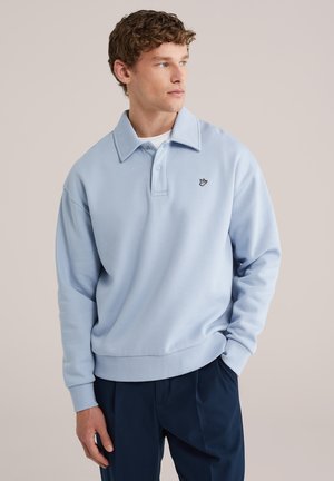 Jonge man met krullend haar, gekleed in een lichtblauwe trui met kraag en een klein logo, donkere broek, met één hand in de zak, kijkt naar rechts.