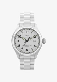 Reloj de pulsera Timex Waterbury plateado con correa metálica, números negros, agujas luminosas, ventana de fecha a las 3, mostrando la hora 10:09:36.