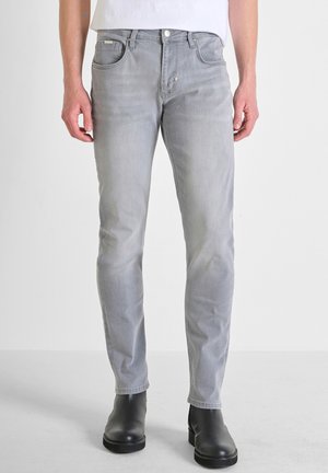 Sivi slim-fit traperici izrađeni od denima, s pet džepova, dugmetom za zatvaranje i blago izblijedjelom bojom. Noseni s crnim čizmama do gležnja.