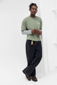 Muškarac s dredovima nosi slojevitu majicu dugih rukava u zelenoj i sivoj boji, crne široke traperice s privjeskom, stoji i naslanja se na bijeli zid.