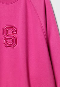 Sweat-shirt rose vif avec une lettre "S" cousue sur la poitrine gauche, doté d'un col rond et d'une couture de manche raglan.