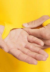 Indumento in tessuto elastico giallo tirato sul manico, che rivela un palmo. La texture è liscia ed elastica, con una vestibilità aderente attorno al polso.