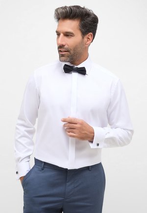 Weißes Hemd mit klassischem Kragen, schwarzer Fliege und Manschettenknöpfen; getragen mit marineblauen Hosen, die einen glatten, polierten Stoff zur Schau stellen.