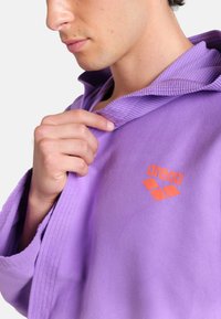 Felpa con cappuccio in pile viola con collo a coste, caratterizzata da un logo arancione. Il tessuto ha una texture morbida ed è leggermente aderente intorno al collo.