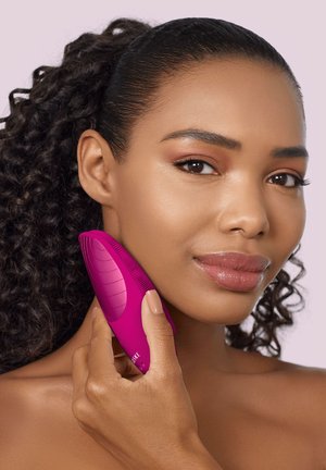 Femme aux cheveux bouclés utilise un appareil de nettoyage du visage rose vif sur sa joue, sur un fond violet pâle.