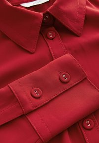 Chemise rouge  à manches longues avec un col pointu, une patte de boutonnage et des poignets boutonnés présentant des détails de couture visibles.