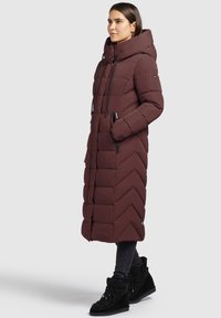 Lange burgundy gewatteerde jas met capuchon, voorzien van chevron-stiksel, zijzakken en zwarte trekkoorden. Draagt met zwarte bont gevoerde enkelboots.