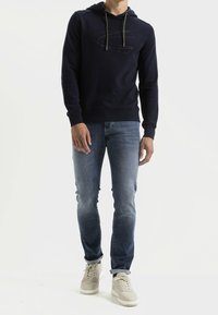 Granatowy hoodie z podszewką w kapturze i detalem logo, w połączeniu z jasnoniebieskimi dżinsami i beżowymi sneakersami, ukazującym swobodny styl.