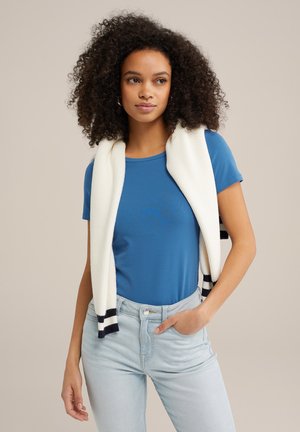 T-shirt bleu à manches courtes sous un cardigan blanc à côtes, présentant des rayures noires et bleu marine aux extrémités, associé à un jean en denim clair.