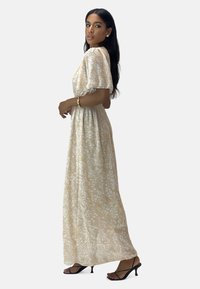 Robe maxi à imprimé floral en tissu beige clair avec des manches courtes et une taille cintrée. Associée à des sandales à talons hauts noires.
