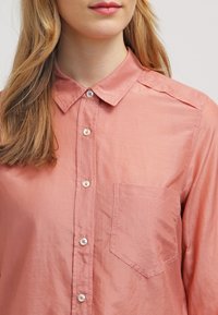 Ljust rosa skjorta med knappar som har en klassisk krage, framficka och slät, något glänsande yta. Traditionell knäppning med knappar.