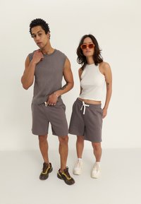Dva modely majú na sebe bežné oblečenie: ribbed šedý top bez rukávov, šedé šortky s bielym šnúrkovým pásom a vzorované tenisky; žena má na sebe biele tielko.