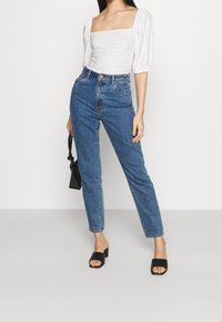 White puff-sleeve top with cut-out details, paired with high-waisted blue jeans. Black open-toe slide sandals. Model holds a small black bag.