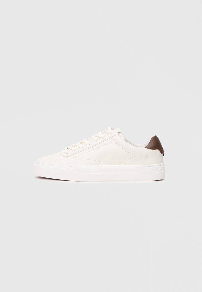White leather sneaker with a smooth texture, round toe, and a brown accent at the heel. Features a lace-up front and thick white sole.