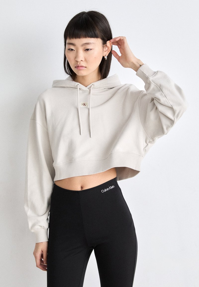 Hellbeige verkürzter Hoodie mit einer Fronttasche, Kapuze mit Kordelzug und geripptem Saum, kombiniert mit hochtaillierten schwarzen Leggings mit einem Logo.