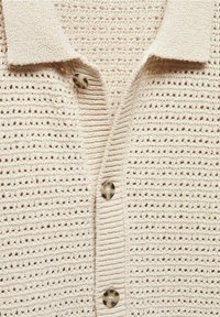 Beżowy sweter robiony na drutach z kołnierzykiem, z teksturowanym wzorem i trzema okrągłymi guzikami z jasnego materiału.