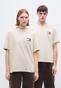 Μπεζ βα μβακερές κοντομάνικες μπλούζες με στρογγυλή λαιμόκοψη, με ένα μικρό λογότυπο σε ναυτικό μπλε, κόκκινο και λευκό στο αριστερό στήθος.