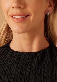 Boucles d'oreilles en argent avec une surface texturée et un léger éclat, portées par une personne à la peau claire et aux cheveux blonds raides, portant un haut noir.