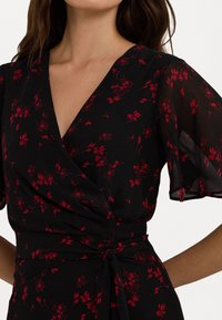 Svart klänning med röd blommönster, korta genomskinliga ärmar, V-ringning och knytning i midjan, tillverkad av lättviktsmaterial.