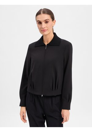 Frau mit glattem Haar, die eine schwarze langärmelige Jacke mit Reißverschluss und schwarze Hose trägt, steht vor einem einfarbigen hellen Hintergrund.