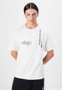 Hvid bomulds t-shirt med et gråt Nike-logo og sort tekst, der læser "International Sportswear" vertikalt langs siden. Korte ærmer.