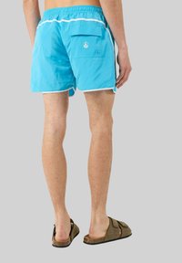 Pantaloni da bagno blu con piping bianco, cintura elastica e una tasca sul retro. Indossati con sandali marroni. Angolo di visualizzazione dal retro.