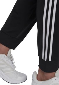 Schwarze Sporthosen mit drei weißen Streifen an den Seiten, kombiniert mit weißen Turnschuhen, die über Mesh- und Gummidetails verfügen.