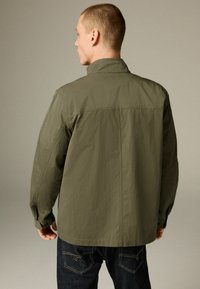 Homme portant une chemise à manches longues olive avec col, de dos, associé à un jean bleu foncé avec des coutures de poches arrière visibles.