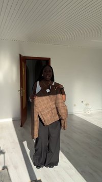 Osoba stojąca w jasnym, pustym pokoju, trzymająca brązową kurtkę w kratę, ubrana w biały top i szerokie czarne spodnie.