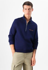 Un giovane con un'espressione seria indossa una felpa con zip blu navy e pantaloni cargo color khaki chiaro, in piedi davanti a uno sfondo neutro.