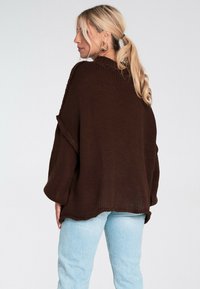 Brązowy sweter dziergany o luźnym kroju, szerokich rękawach i prążkowanym kołnierzu. Noszony na jasnoniebieskich dżinsach o fakturowanej powierzchni.