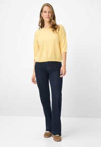 Pull léger jaune à manches trois-quarts, associé à un jean évasé bleu foncé et des chaussures en daim marron, posé sur un fond blanc.