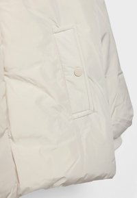 Veste matelassée beige clair avec une surface douce et texturée, fermeture à boutons-pression, et un design épuré et minimaliste. Comprend une poche latérale.