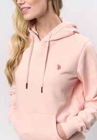 Svetlo roza jopa iz mehkega blaga, s sprednjim žepom, kapuco z vrvico in majhnim vezenim logotipom. Teksturirani manšete in spodnji rob.