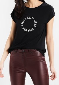 Czarna koszulka z krótkim rękawem i okrągłym dekoltem, ozdobiona białym okrągłym nadrukiem z napisem "Calvin Klein Jeans Nowy Jork". Połączona z bordowymi dopasowanymi spodniami.