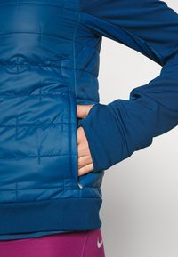 Veste isolante bleue avec un design matelassé, poche latérale et poignets de manches bleus contrastants. Matière lisse avec une finition texturée.