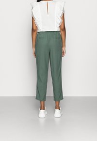 Blusa blanca con mangas acampanadas, detalle de llave en la espalda; pantalones verdes con cintura elástica; la modelo lleva zapatillas blancas.