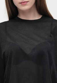 Kvinne iført en gjennomsiktig svart topp som avslører et mørkt undertøy under, med langt brunt hår og synlige naturlige lepper.