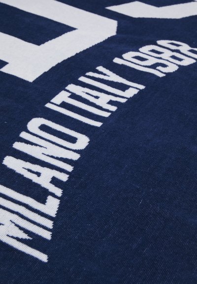 Serviette marine avec des lettres blanches. Le texte indique "MILANO ITALY 1988." Tissu doux et texturé avec un design graphique audacieux.