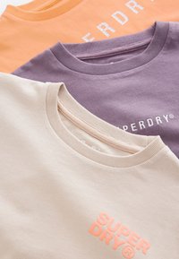 Tre bomull t-shirts i persika, lavendel och krämfärgad. Varje t-shirt har en rund hals och "SUPERDRY"-logotyp i kontrasterande färger.