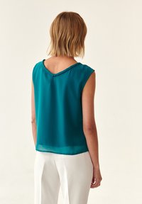 Blusa sin mangas de color verde azulado con escote en forma de V y borde trenzado en el cuello. Tejido ligero con una caída fluida, combinada con pantalones blancos.