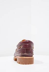 Bateau en cuir marron foncé, avec une semelle robuste et texturée, des œillets dorés et des coutures décoratives le long du dessus et des côtés.